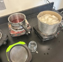 Two pots sit on burners, one containing red liquid, and one containing white wool.