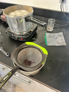 A strainer strains cochineal extract into a pot. Behind it, undyed roving cooks in a pot with mordant.