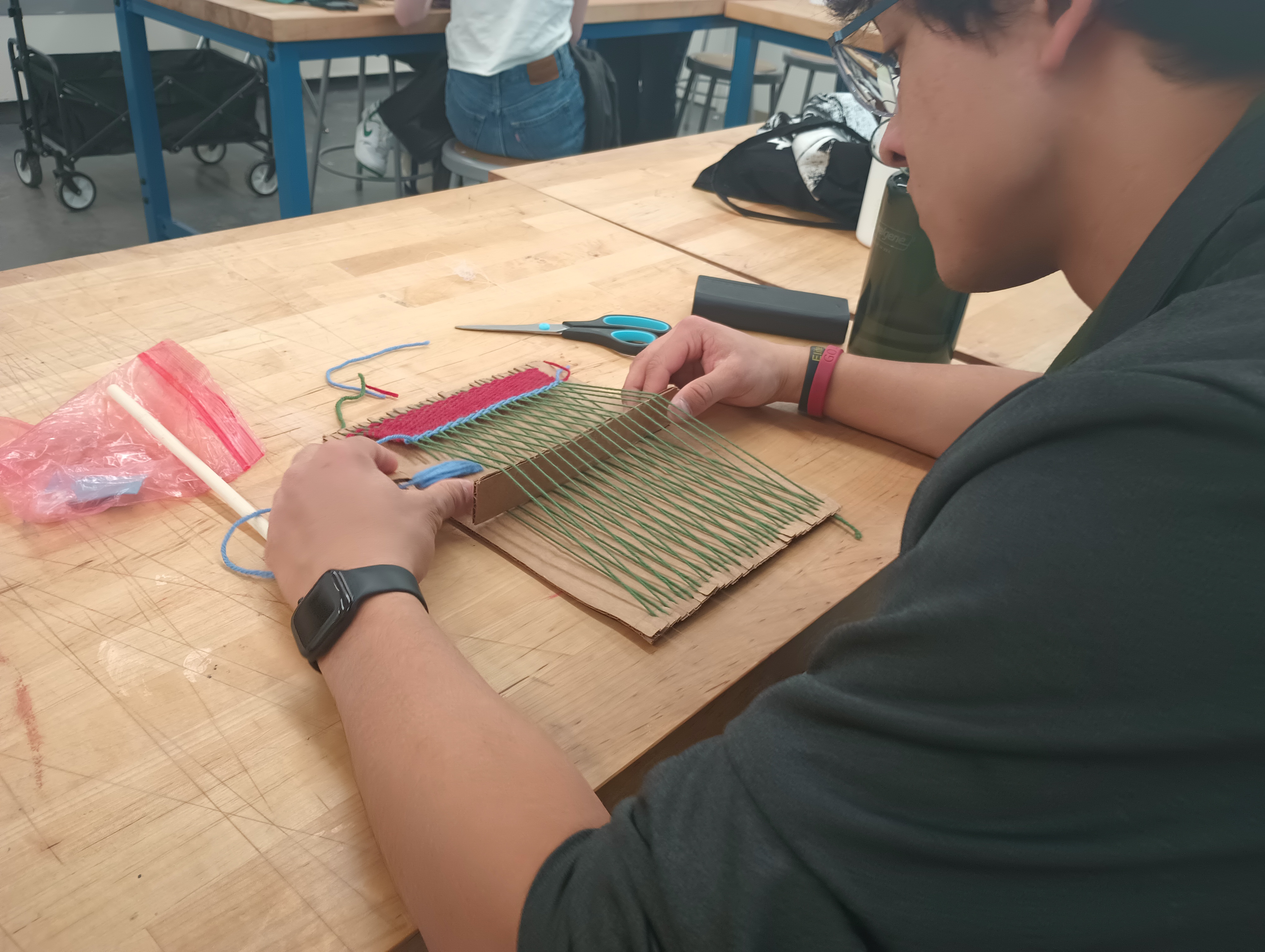A student turns his cardboard shed bar on its side to open up a shed on his cardboard loom. He weaves red and blue wefts into a green warp.