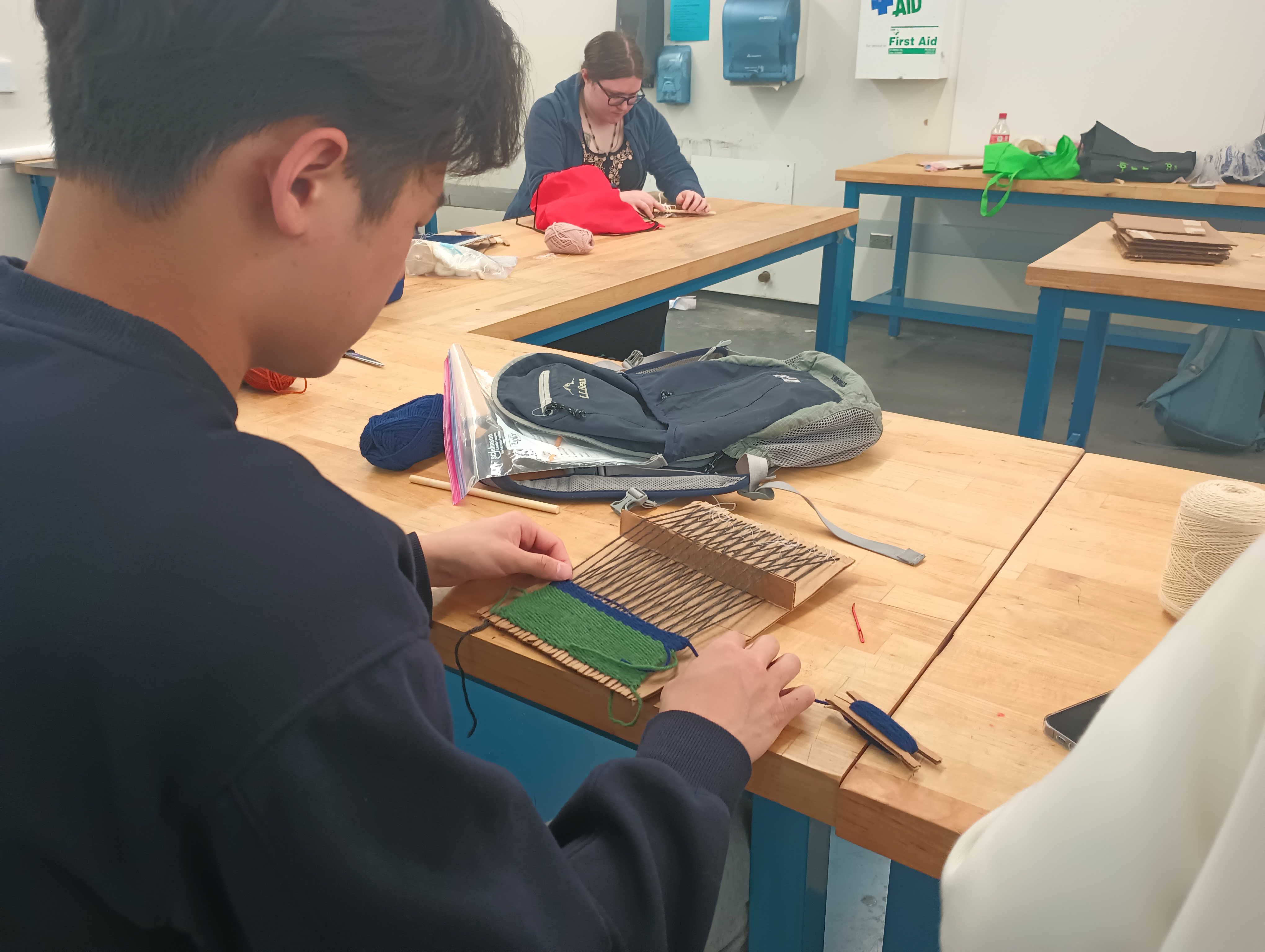 A student evens out a blue weft thread on his cardboard loom. He is adding a blue stripe to a green piece and his cardboard shed bar stands on its side, holding open the shed.