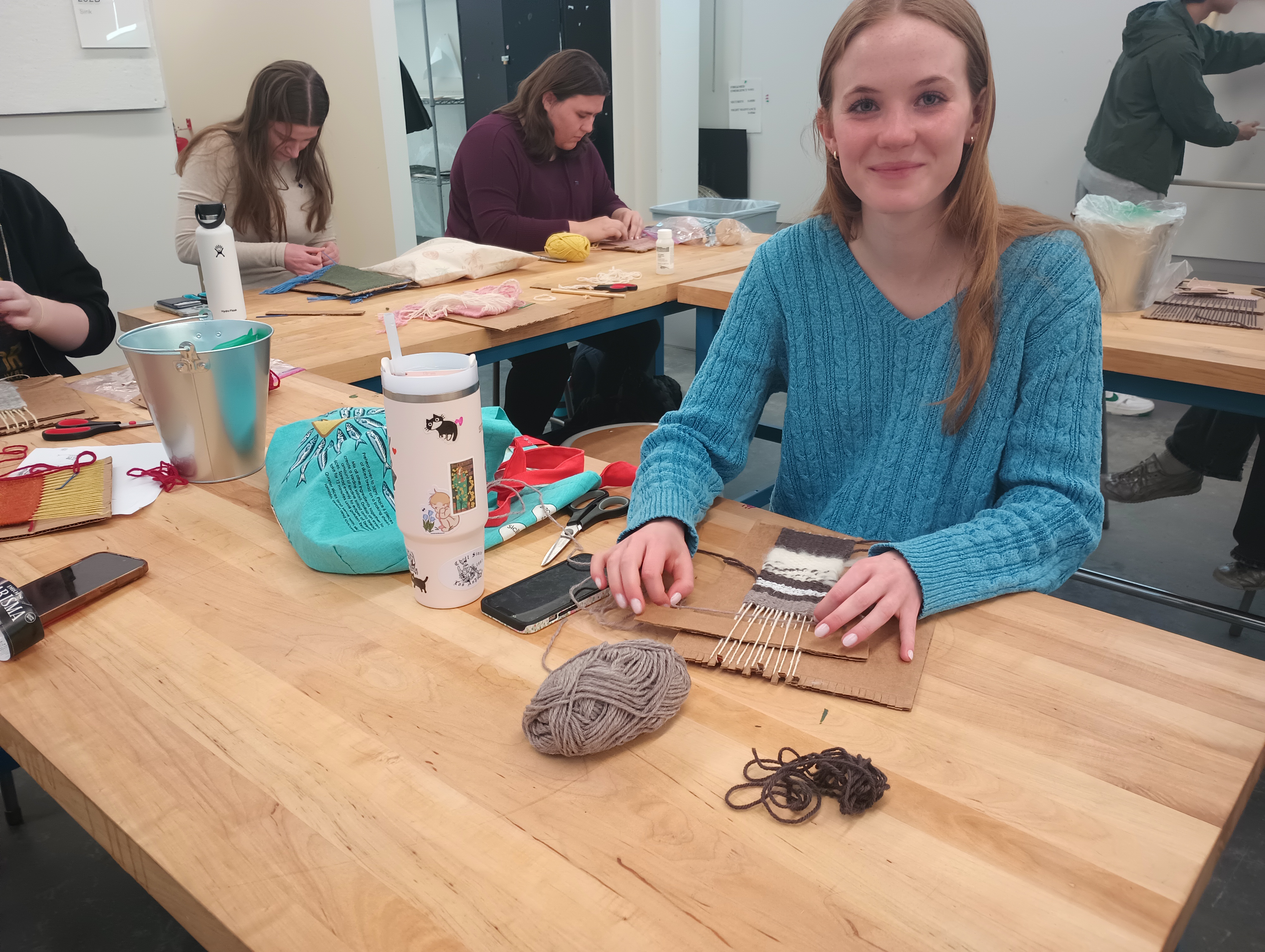A student smiles at the camera as she works on her cardboard loom. She has been weaving a narrow rectangle with stripes of white, brown, and grey yarn. The white weft yarn she spun herself. 