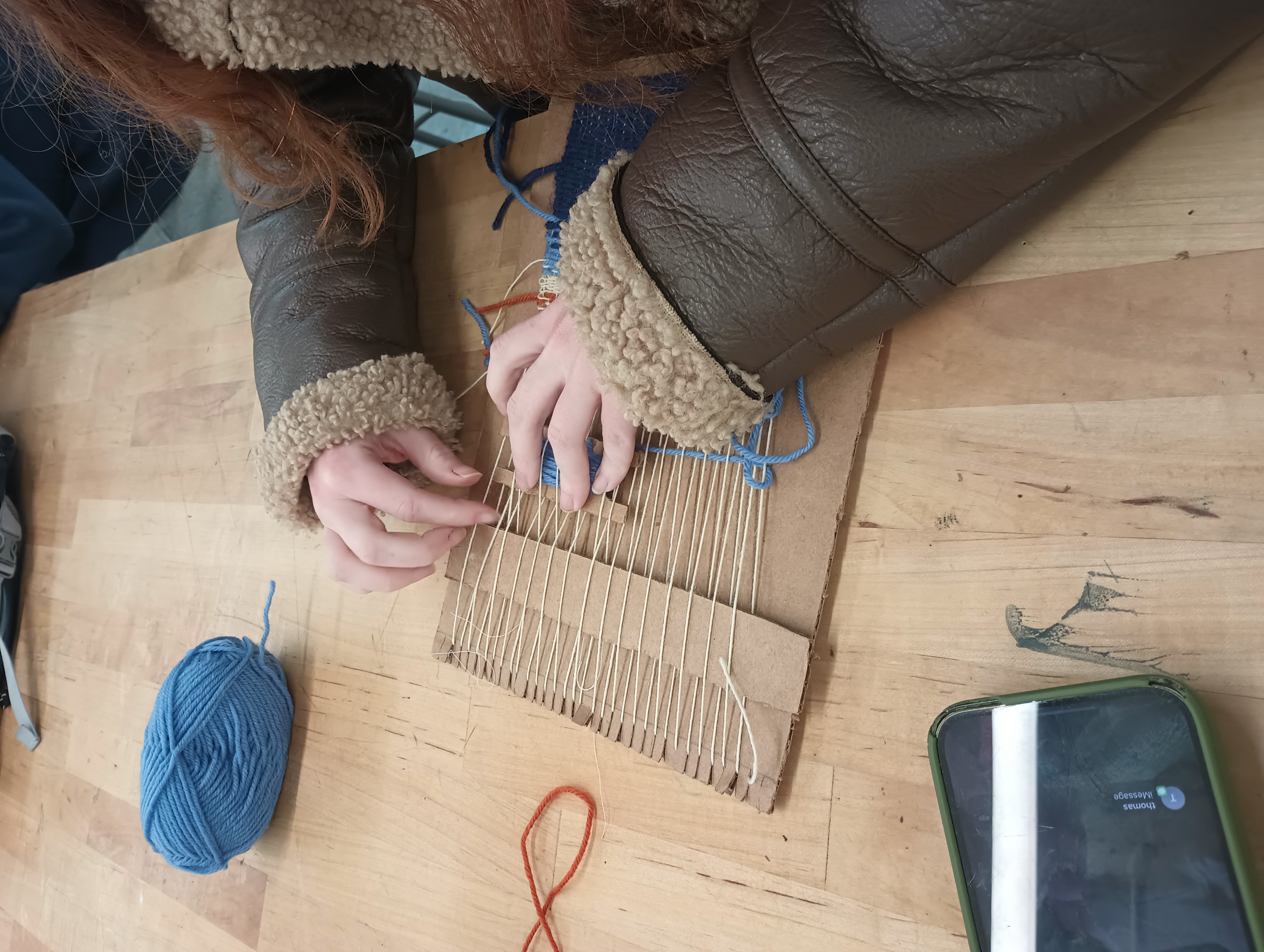 A student passes a cardboard shuttle with blue yarn through the warp on her cardboard loom by hand.