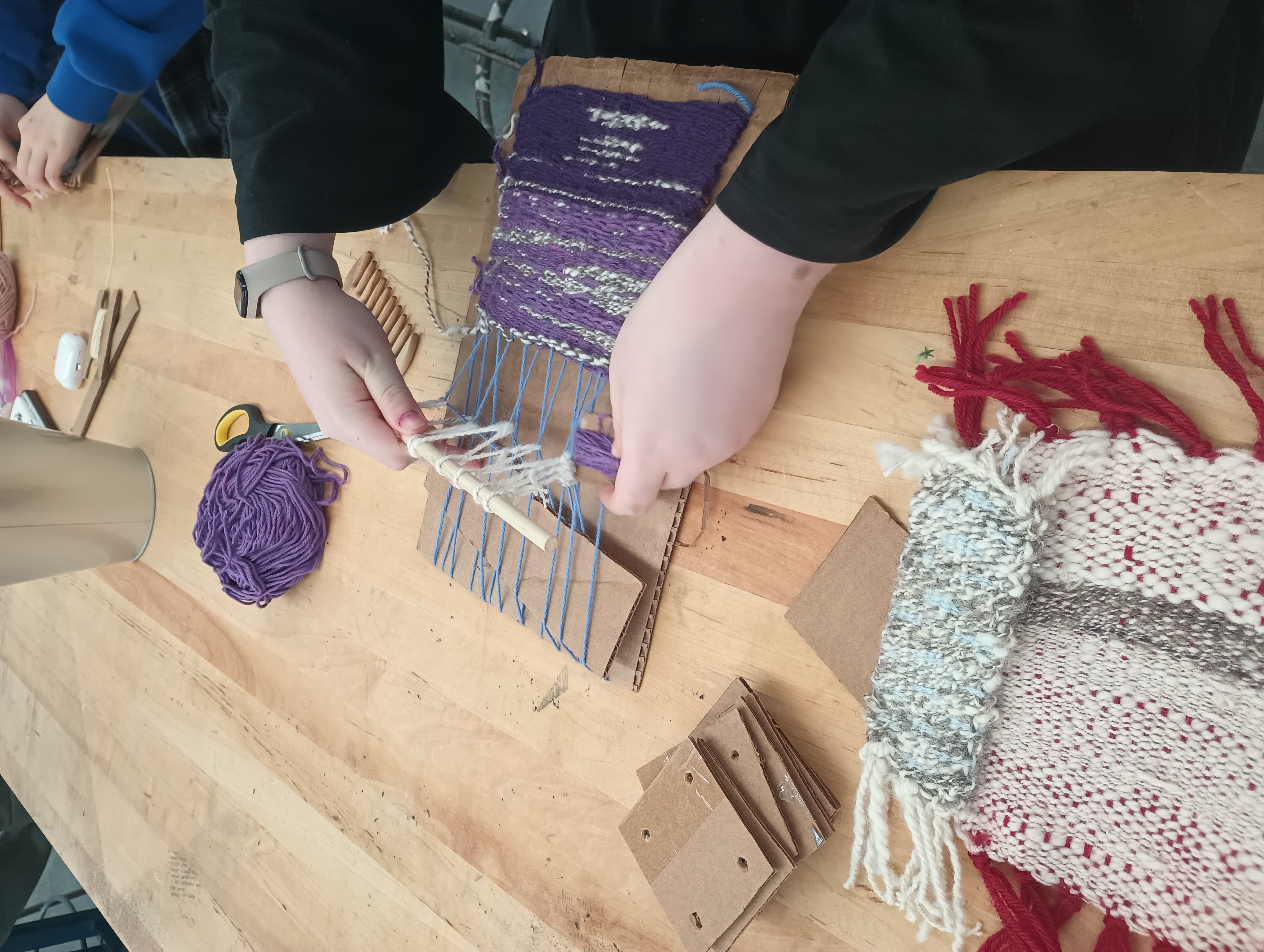 A student lifts a heddle bar and inserts a bobbin of yarn into her piece on a cardboard loom. The piece is woven in various shades of purple and a white/grey 2-ply hand-spun yarn. Beside her work is a stack of cardboard tablets and two completed woven pieces. 