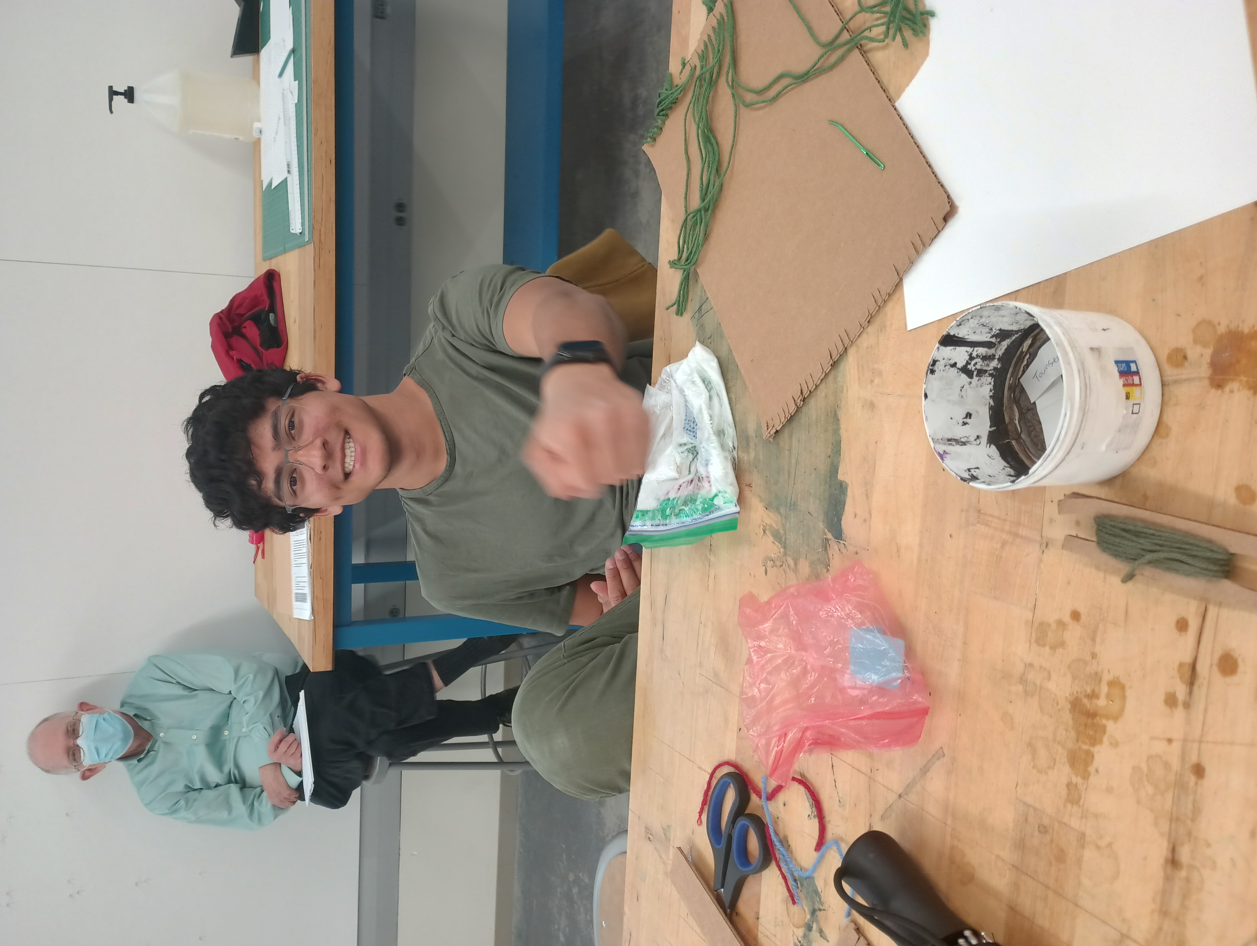 A student smiles at the camera while pounding a plastic bag containing wool, soap, and water with his fist. Behind him smiles classics department chair Timothy Moore, observing this week's studio.
