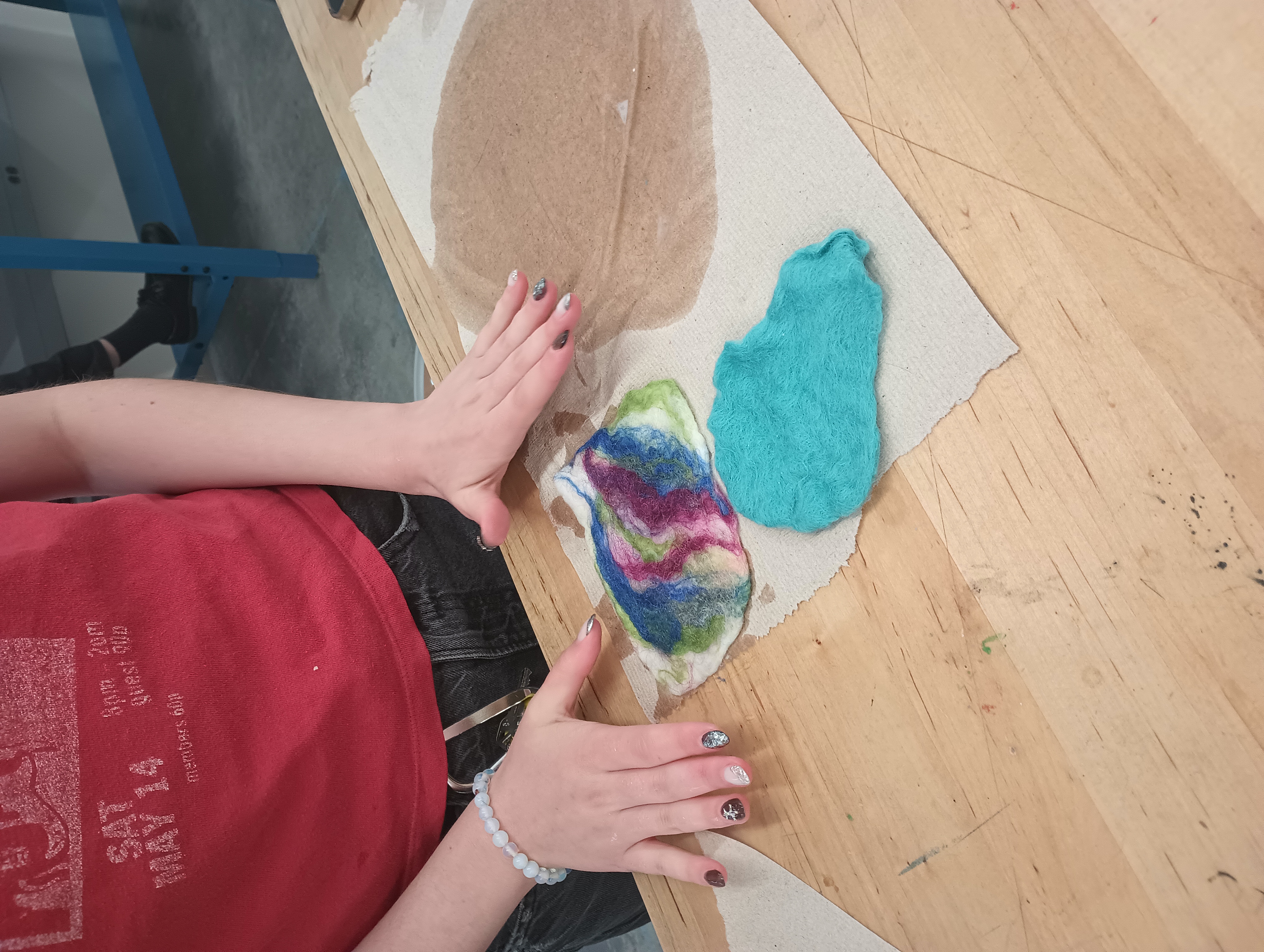 A student lays out a sheet of felt with a white background and bright colorful stripes artfully arranged. Next to thsi piece is a previously completed thin sheet of blue felt.