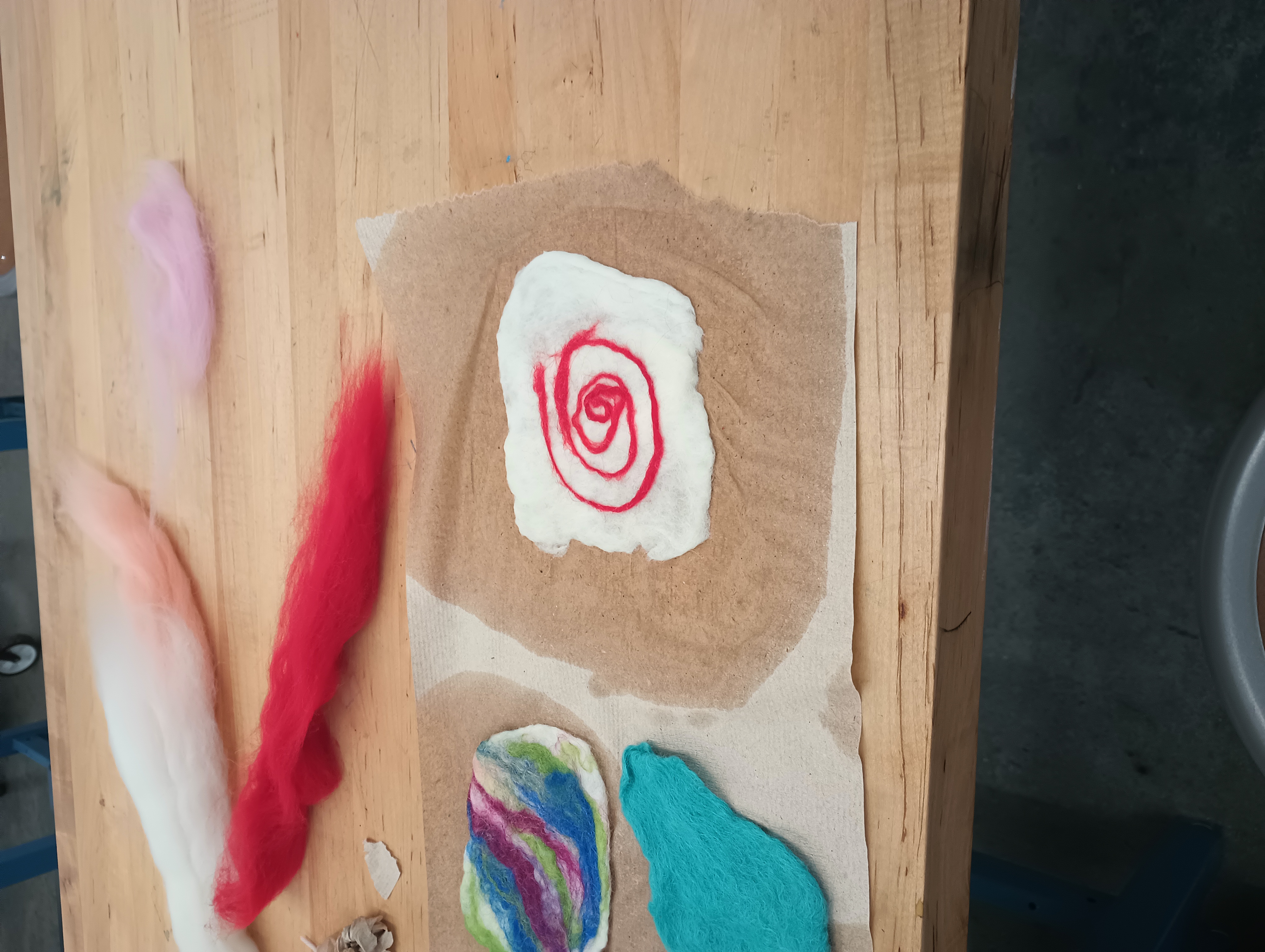 Several pieces of colorful felt drying on a paper towel with some colored unfelted wool roving behind them. One piece is white with a red spiral in the center. 