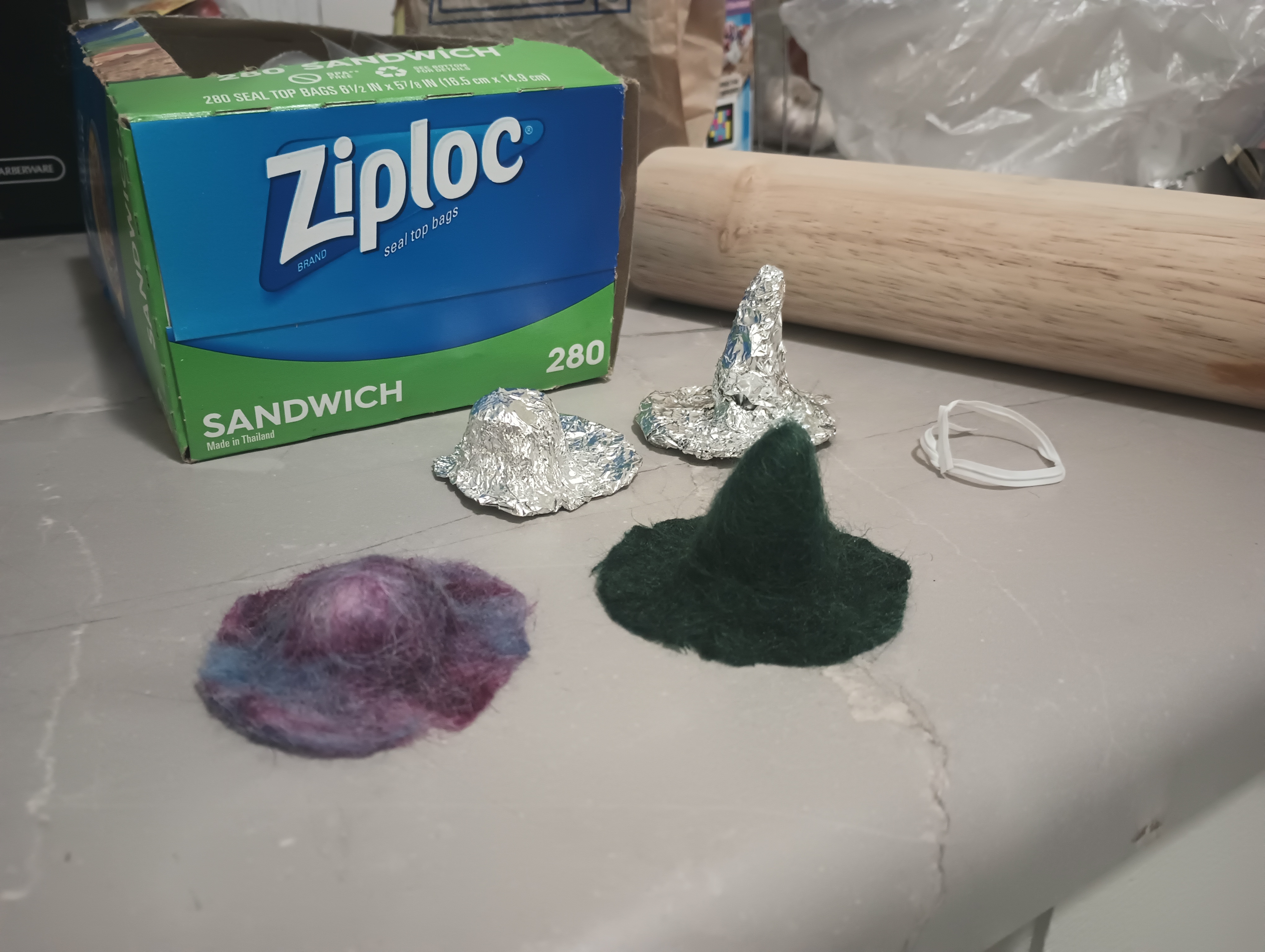 A miniature felted purple hat and a miniature felted dark green witch's hat. Behind them sit tinfoil forms used to shape the hats.