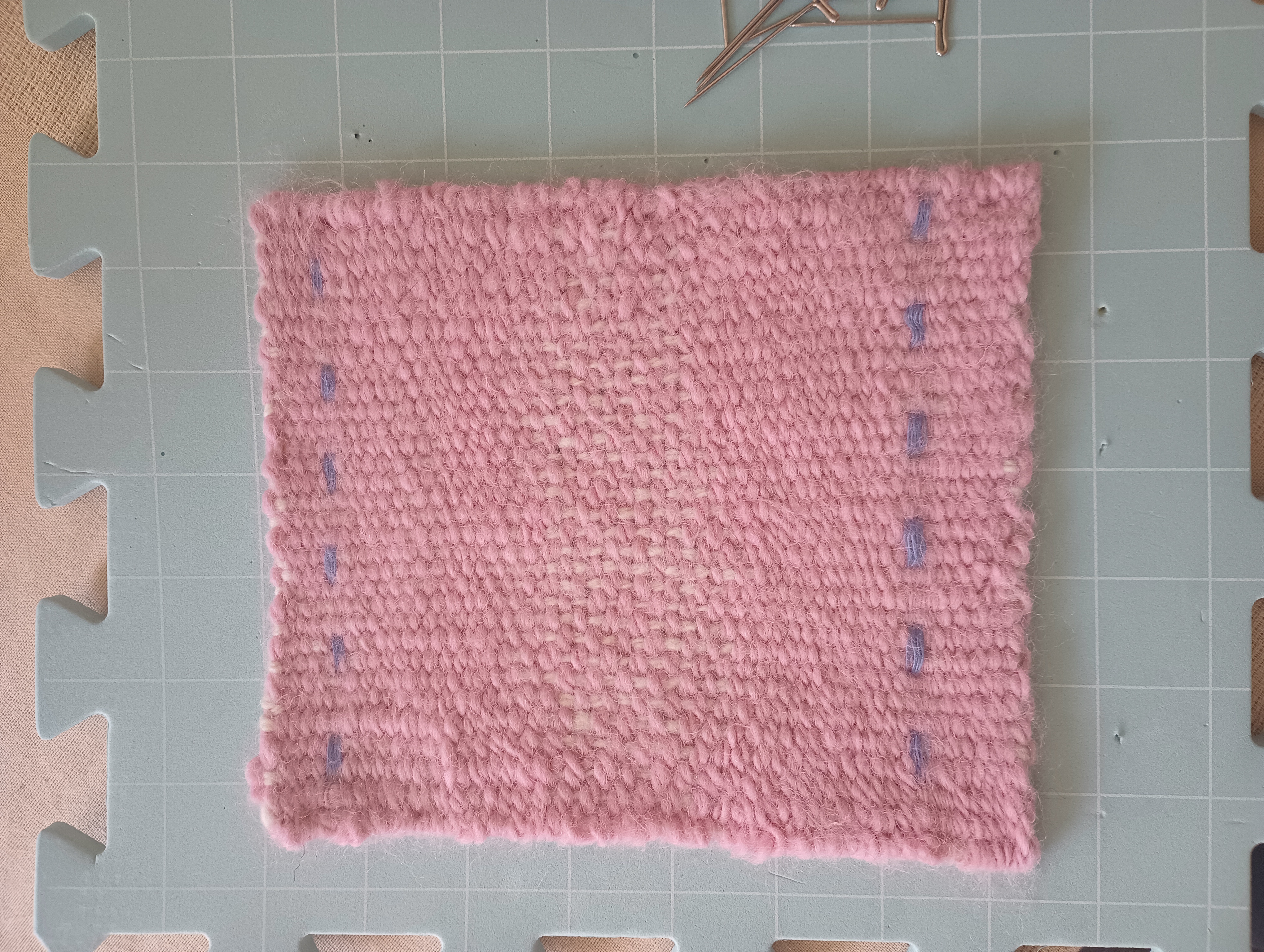 A woven rectangle of white warp, pink weft, and with some blue checks. The tabby weave is quite evident, especially where it is balanced instead of weft-faced, but the texture of the fabric is much fuller and denser looking than before fulling.