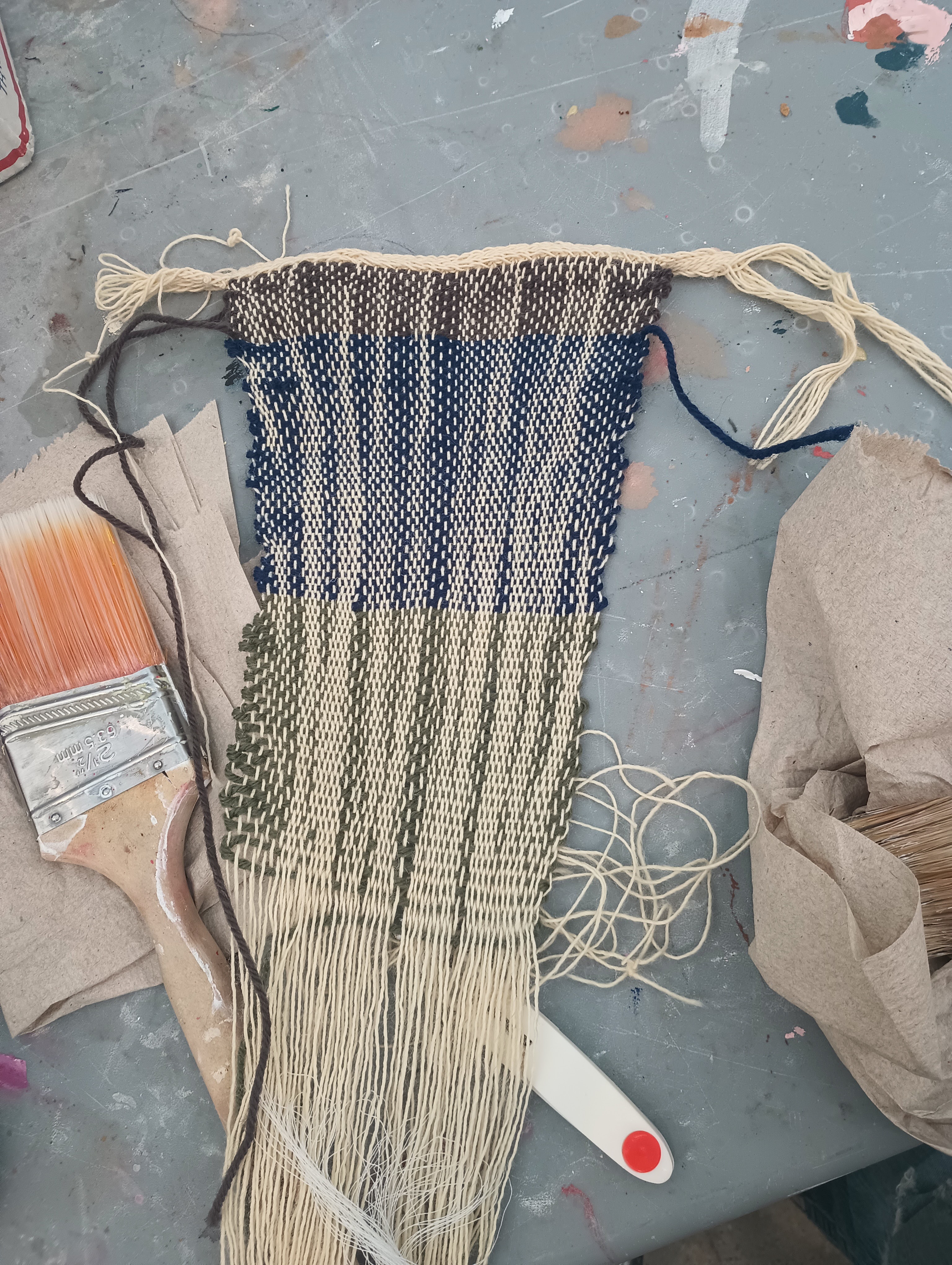The finished piece of cloth woven collectively by students on the warp weighted loom. The cloth has visible white warp threads. The weft alternated between sections of green and blue. The edges are a bit messy and the piece narrows as it gets longer.