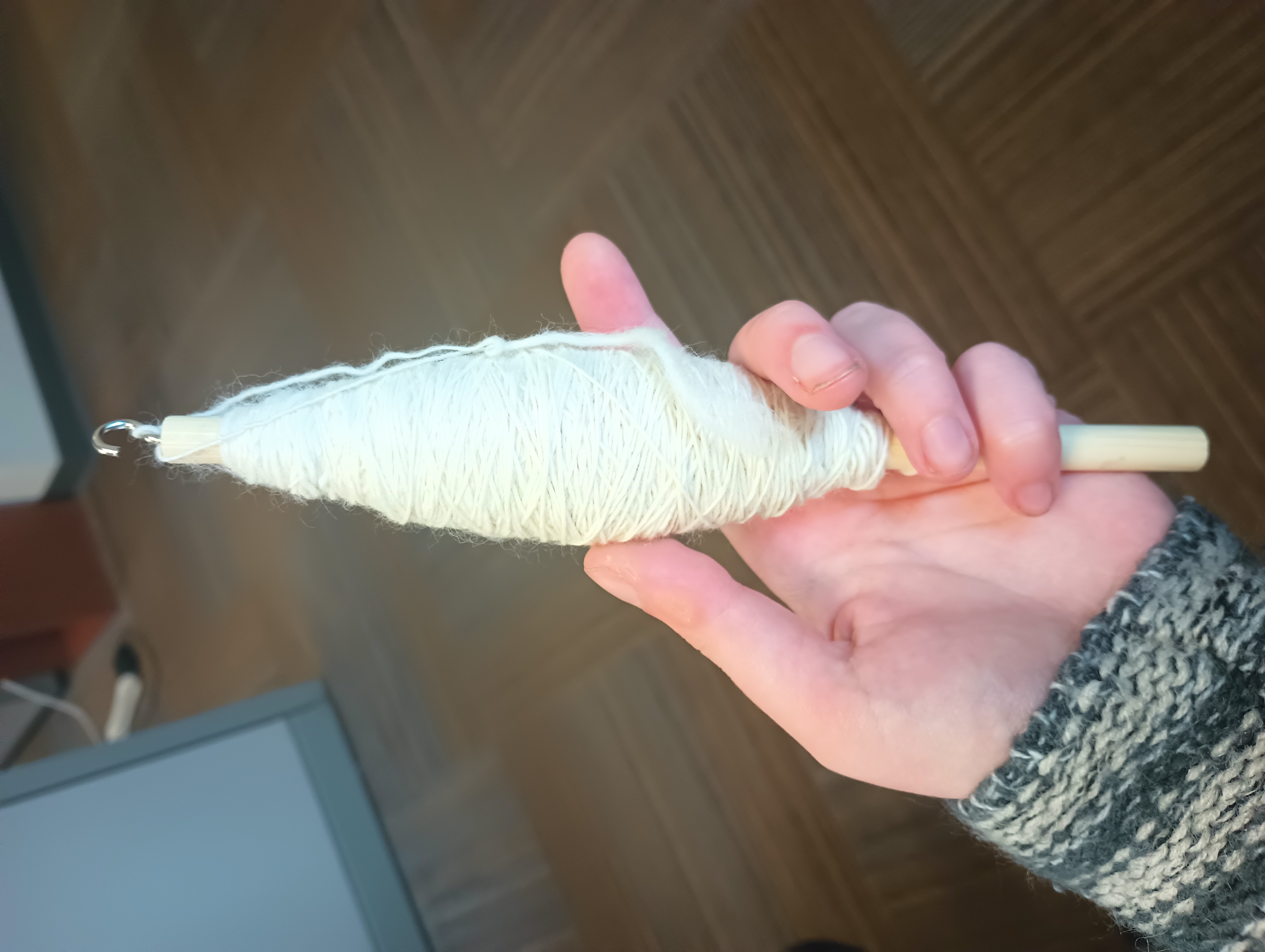 My hand, holding a wooden dowel with a hook on the end wound with a cop of white hand-spun yarn.