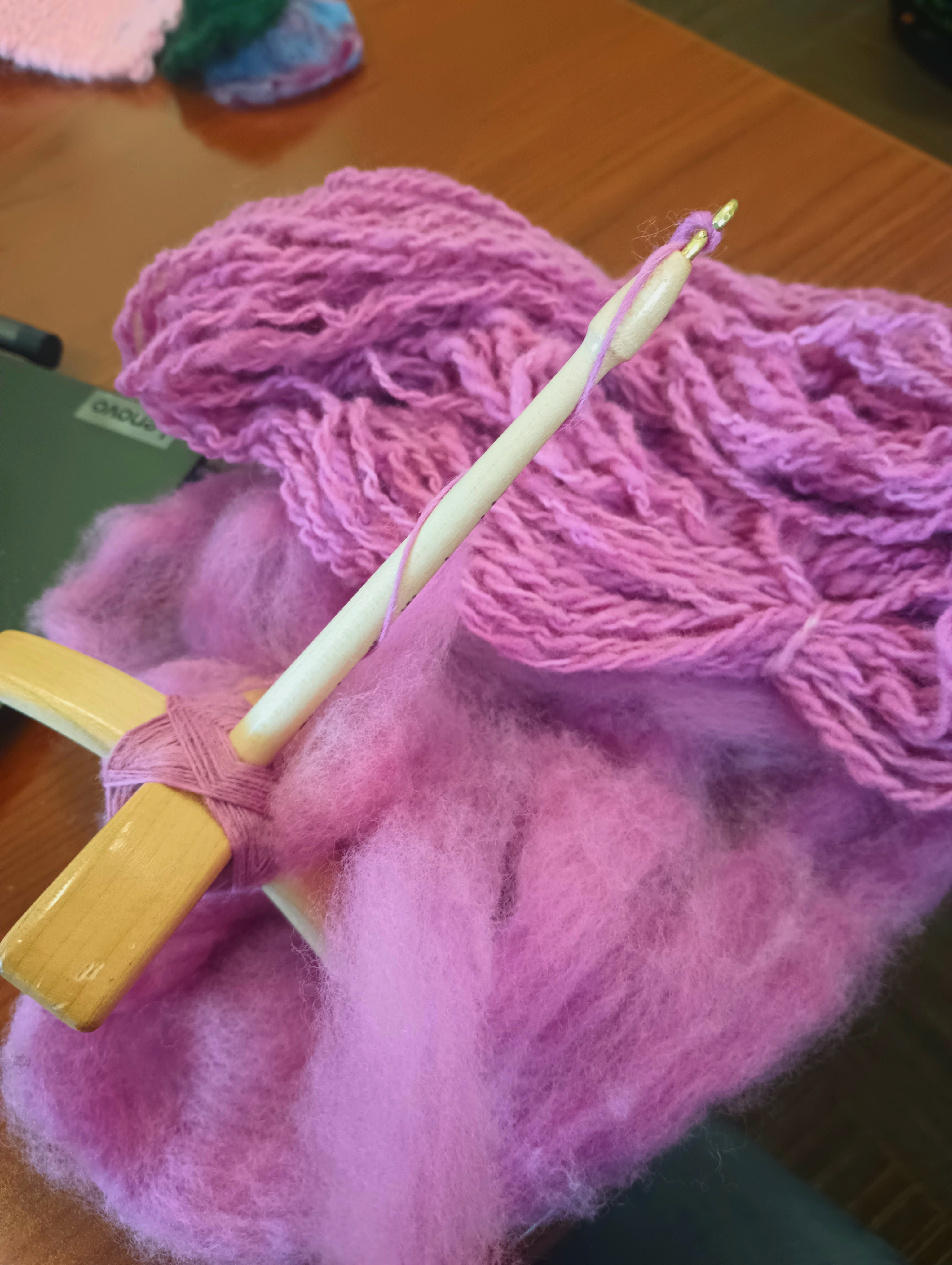 A turkish spindle sits on a pile of fluffy purple-pink roving. There is some spun yarn wound onto the spindle, and also some dyed yarn of the same color in the background.