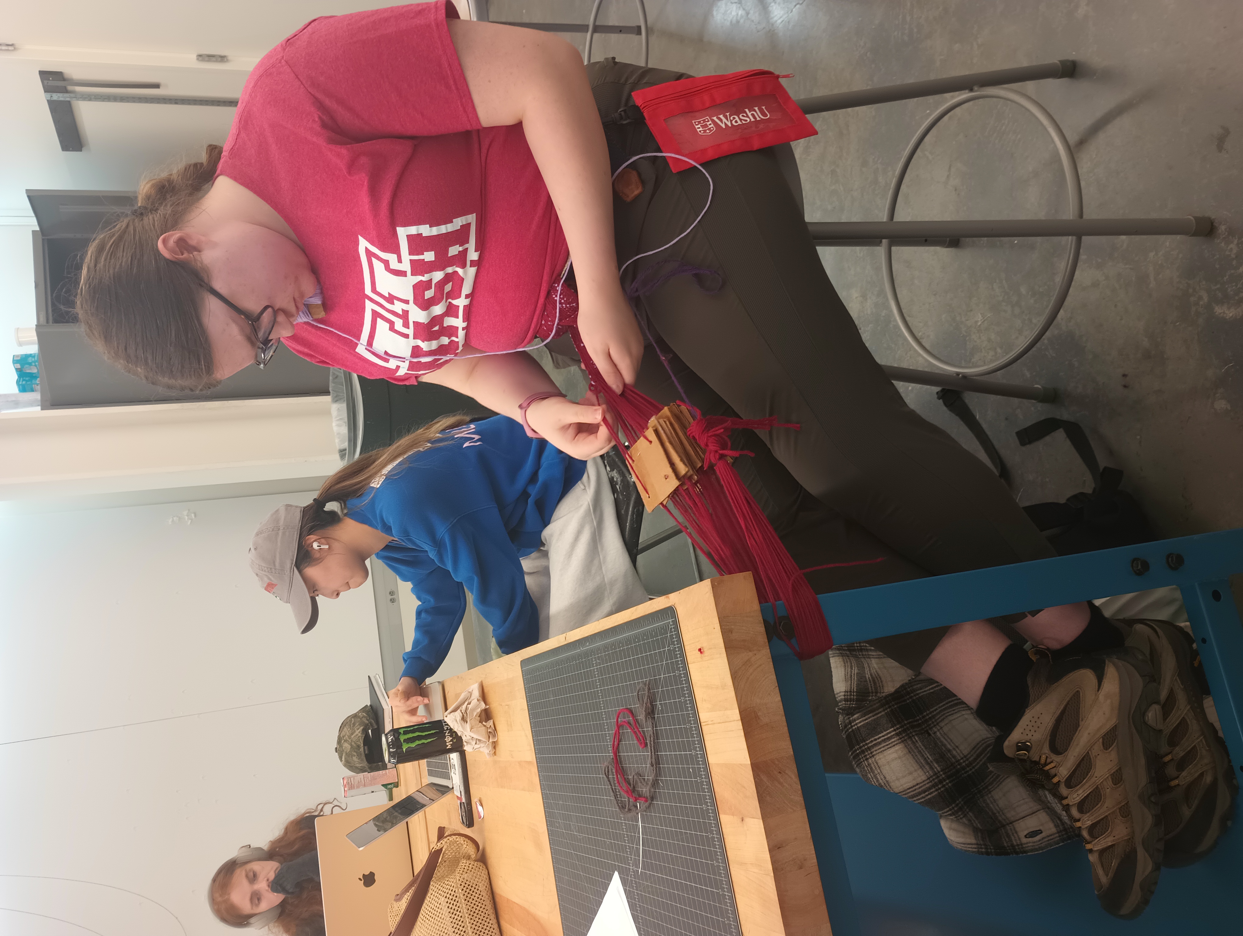 A student works on a tablet weaving project. One end of her red warp is tied to a belt around her waist, and the other to a table leg. A stack of cardboard tablets are threaded onto the warp and she passes the weft through the open shed created by the tablets.