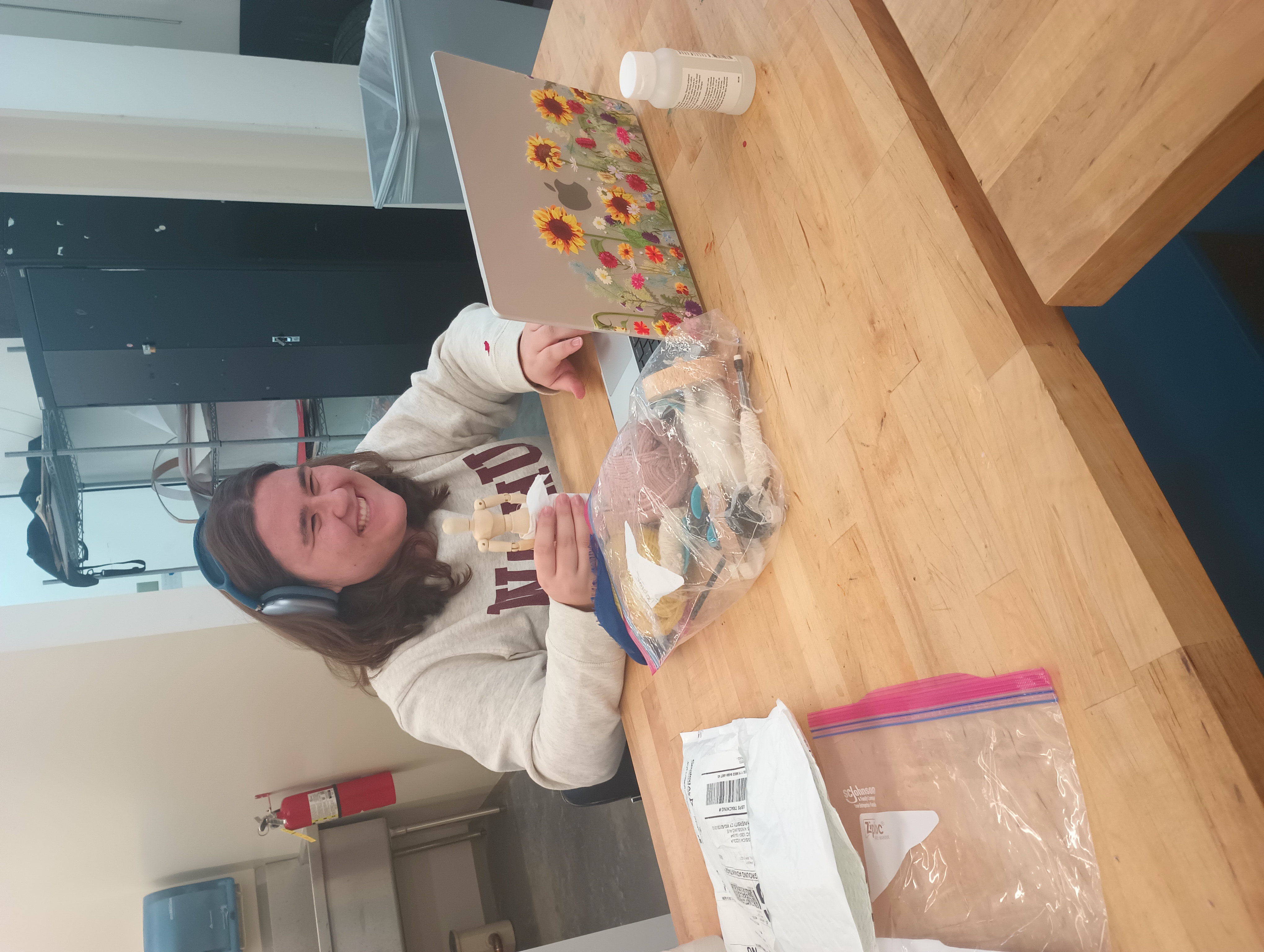 A student holds a small art mannequin and smiles as she works on her laptop. In front of her is a pack of supplies for constructing miniature Greek outfits.