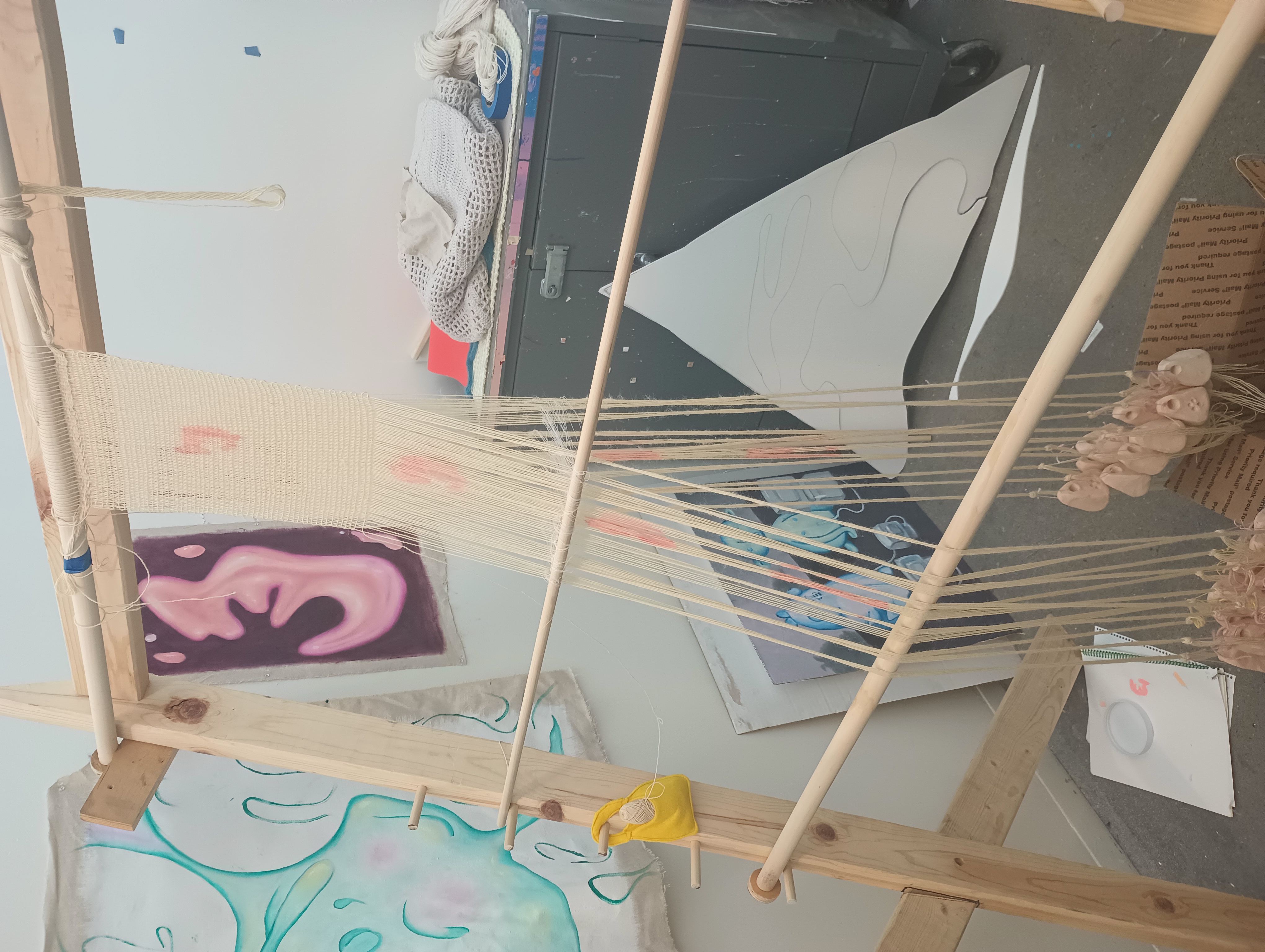 A piece of weaving in progress on the warp-weighted loom. white warp threads are painted with a pale pink design which appears striated as the white, loosely packed weft passes through.