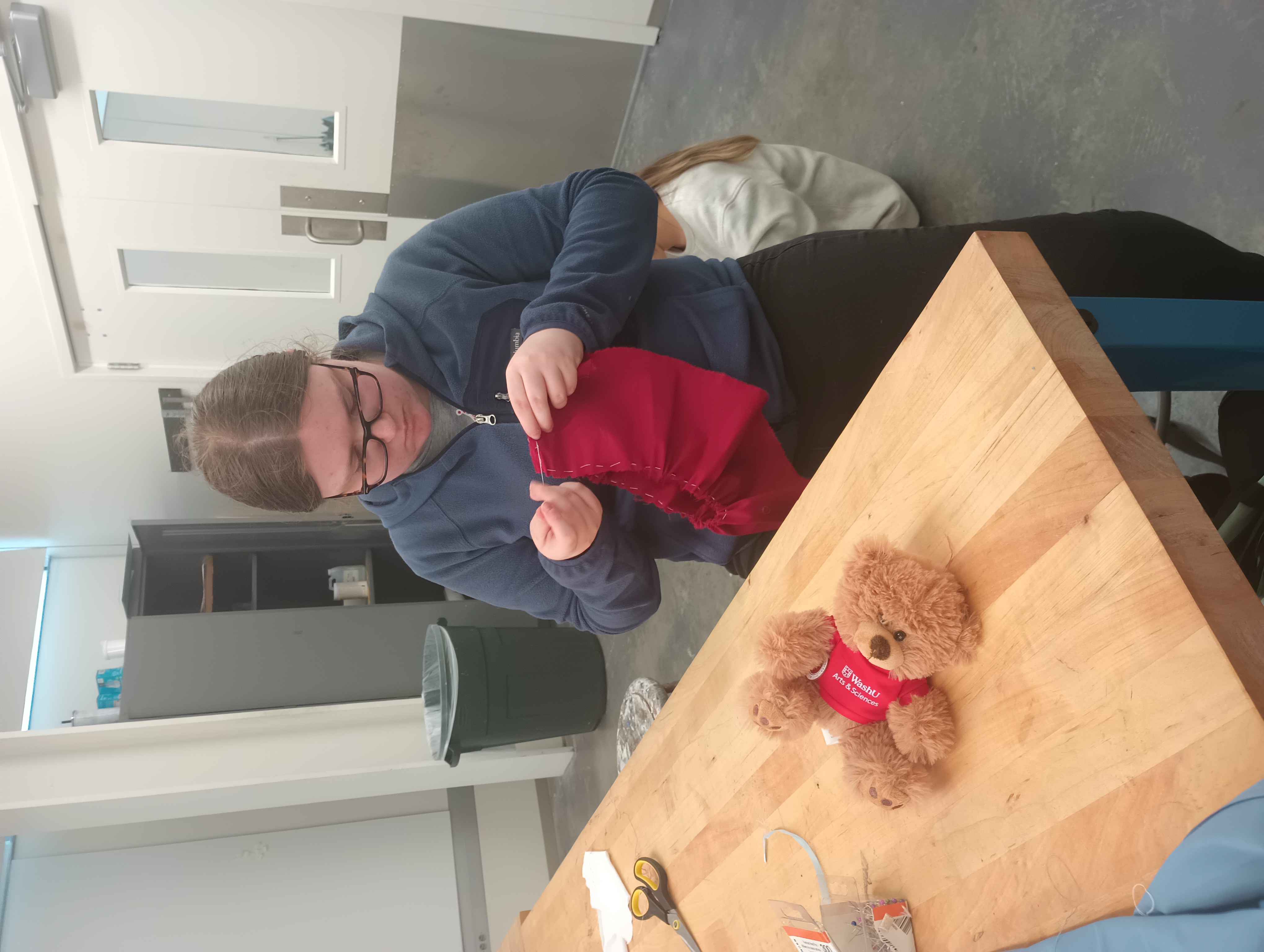 A student sews a line of running stitch around the top of a circle of red fabric, preparing to gather it into a ruffled skirt.
