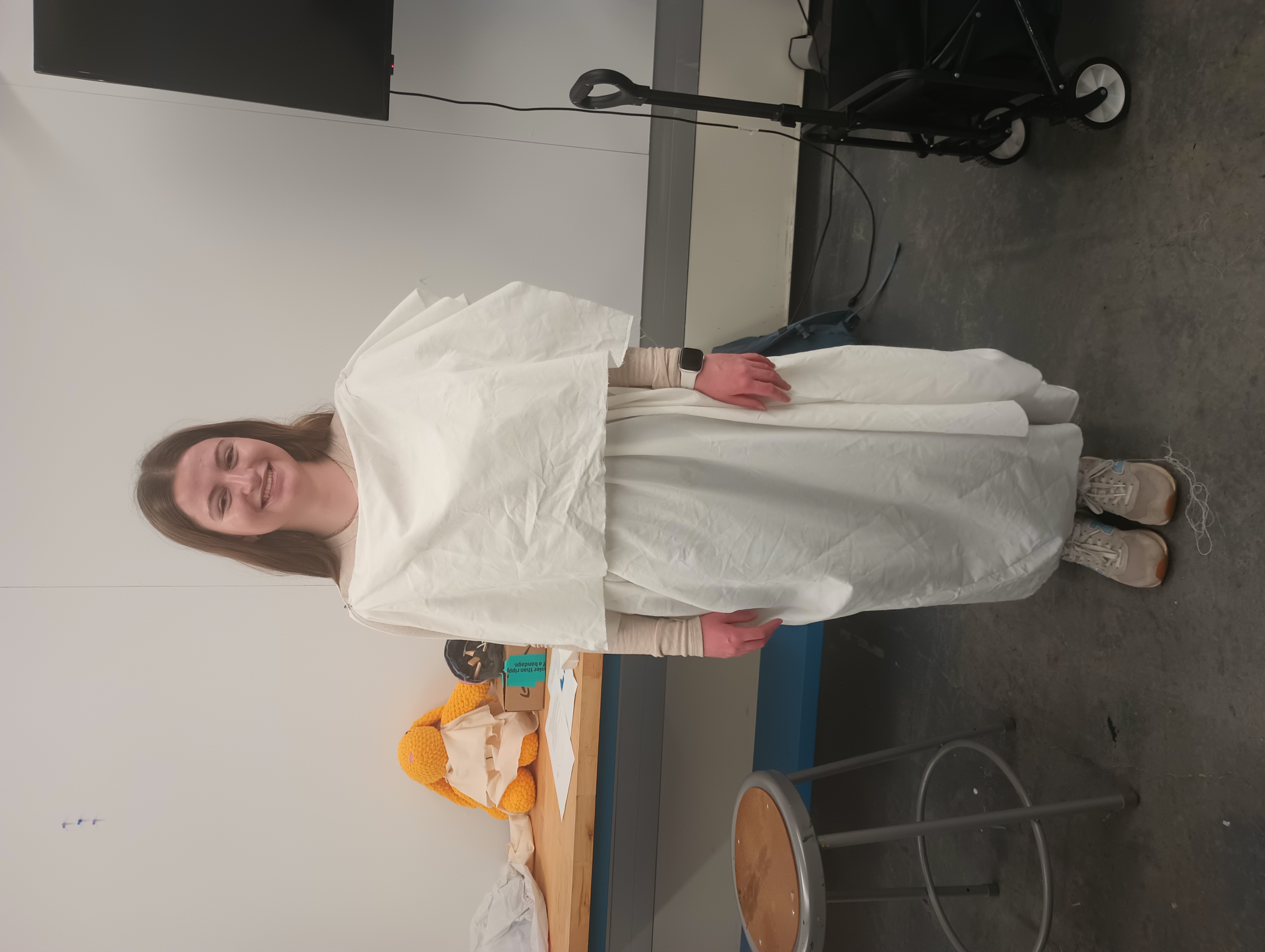 A student wearing an ancient Greek peplos fashioned out of a large white piece of fabric. Behoind her on a table is a stuffed rabbit also wearing a white peplos. 