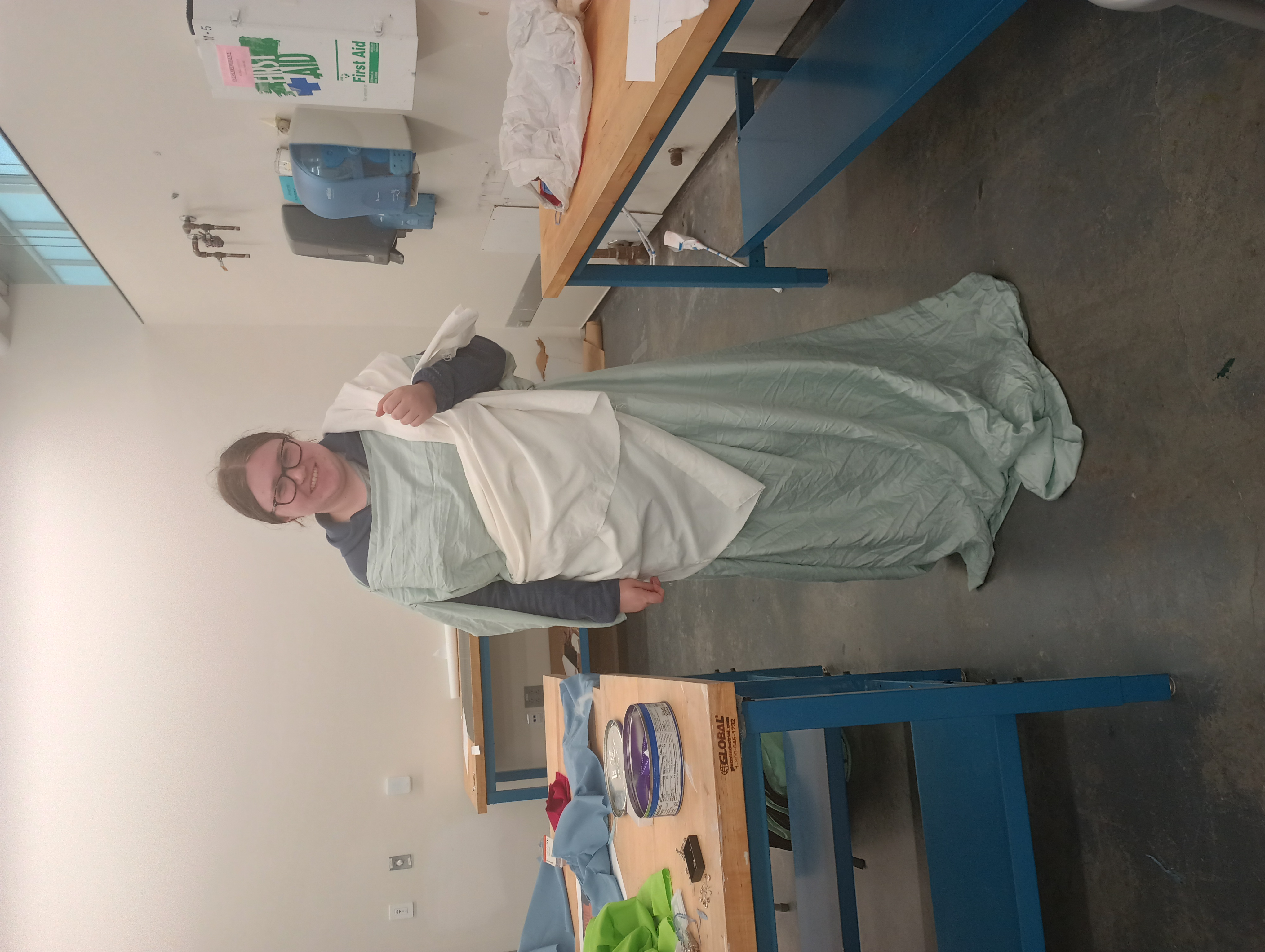 A student wearing a long chiton fashioned from a green sheet and a small white himation draped over a shoulder and one arm. 