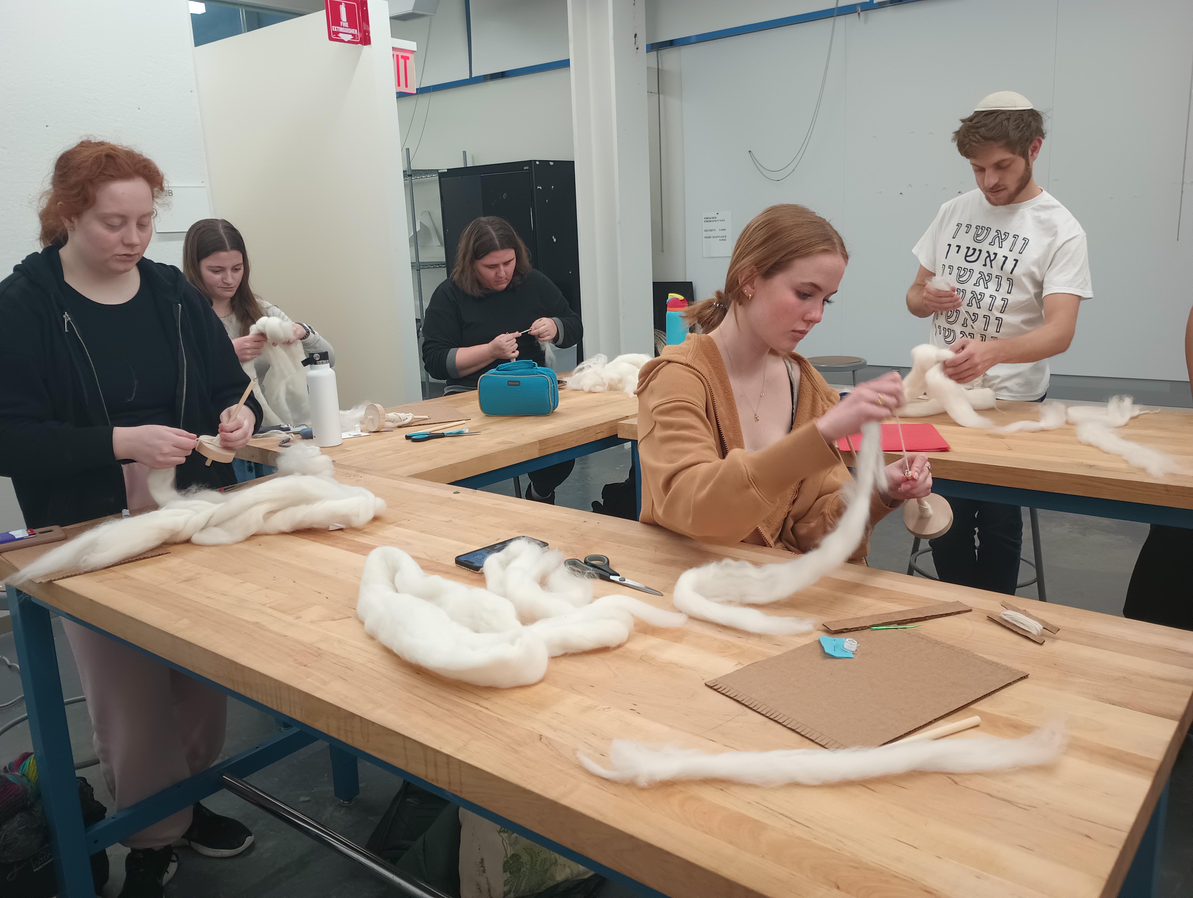 Five students spinning white wool roving into yarn on wooden bottom-whorl drop-spindles. 