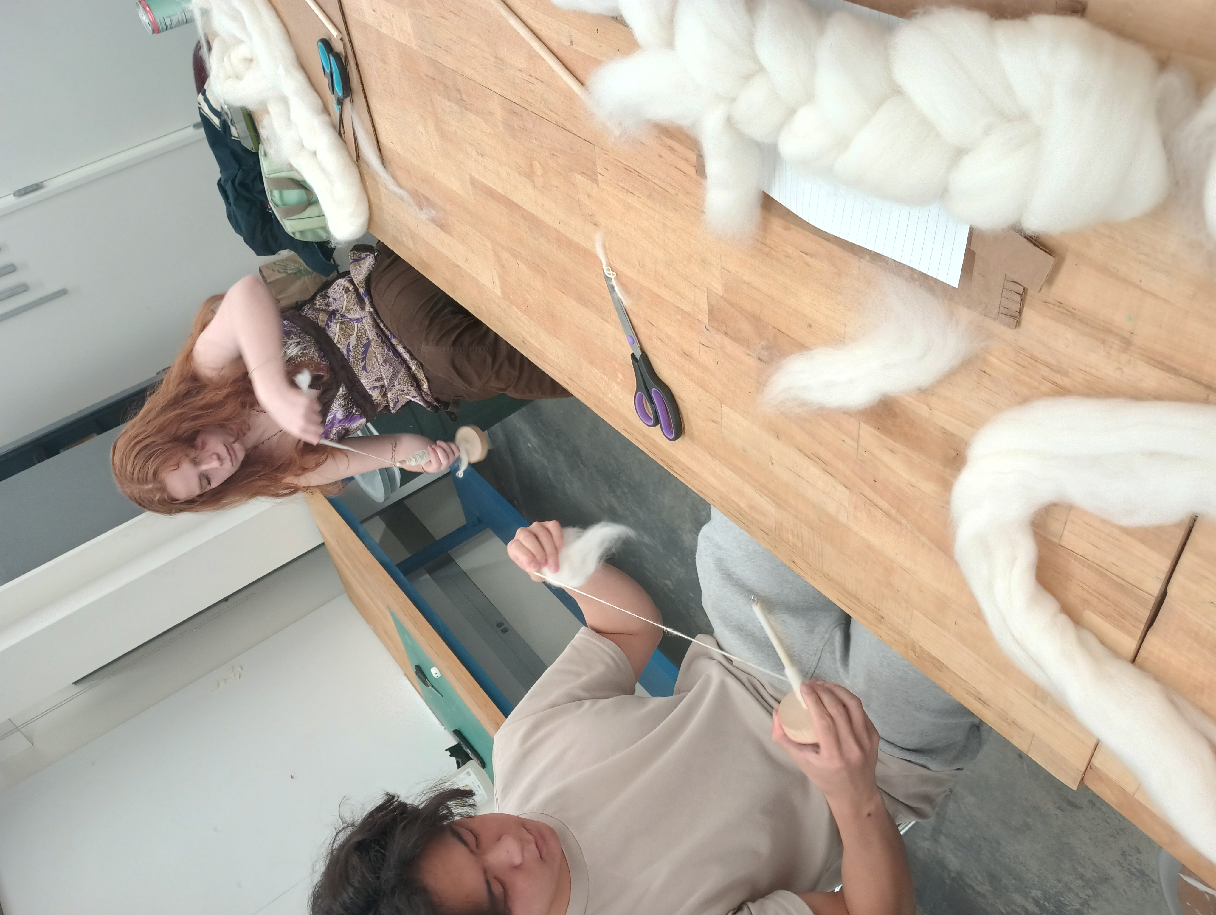 Two students sit at a table spinning. One winds spun yarn onto the shaft on his spindle, while another drafts fiber.