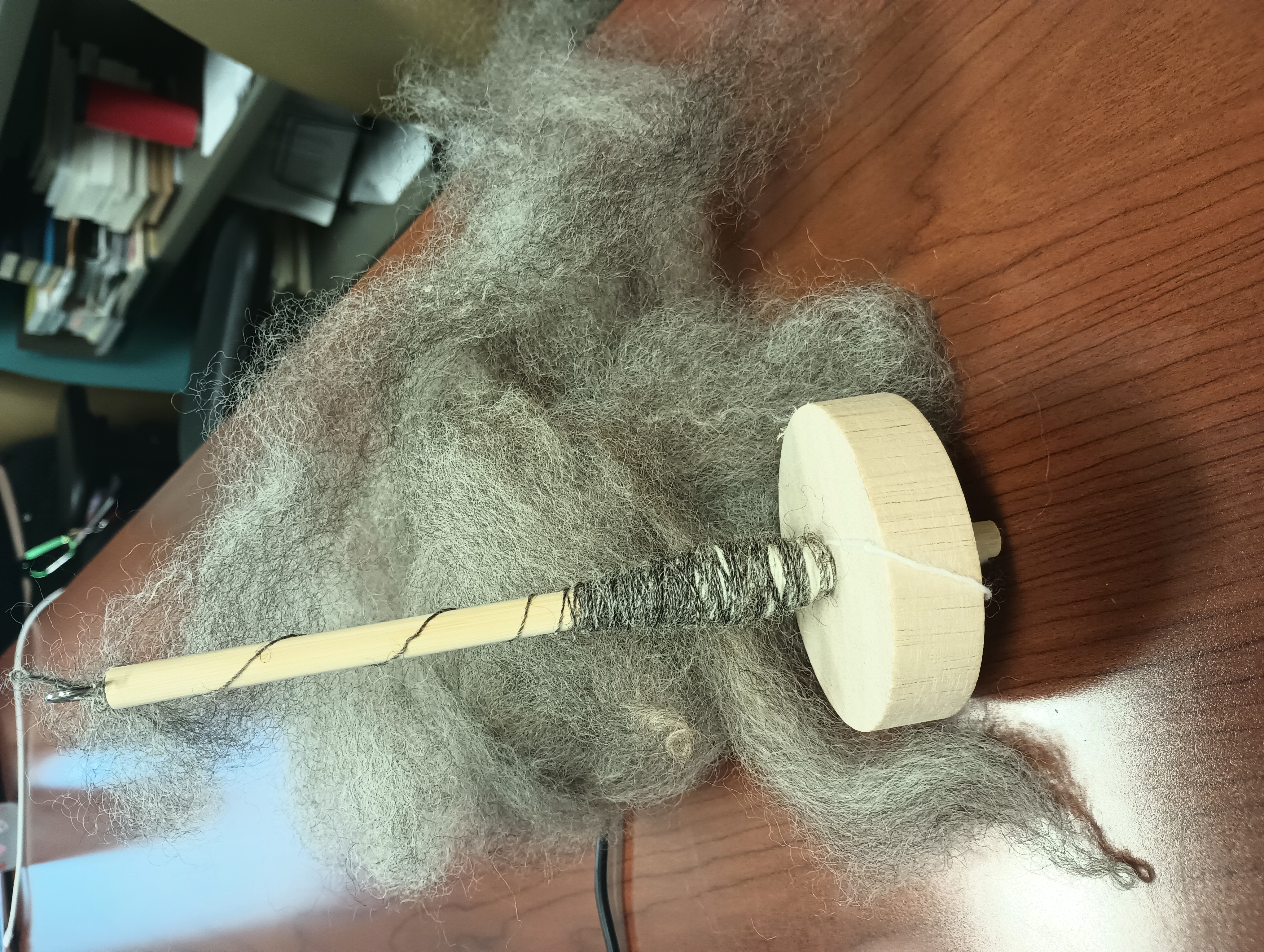 A wooden bottom-whorl drop spindle leans against a fluffy mass of gray wool. A small amount of grey thread is wound onto the spindle and connected to the wool.