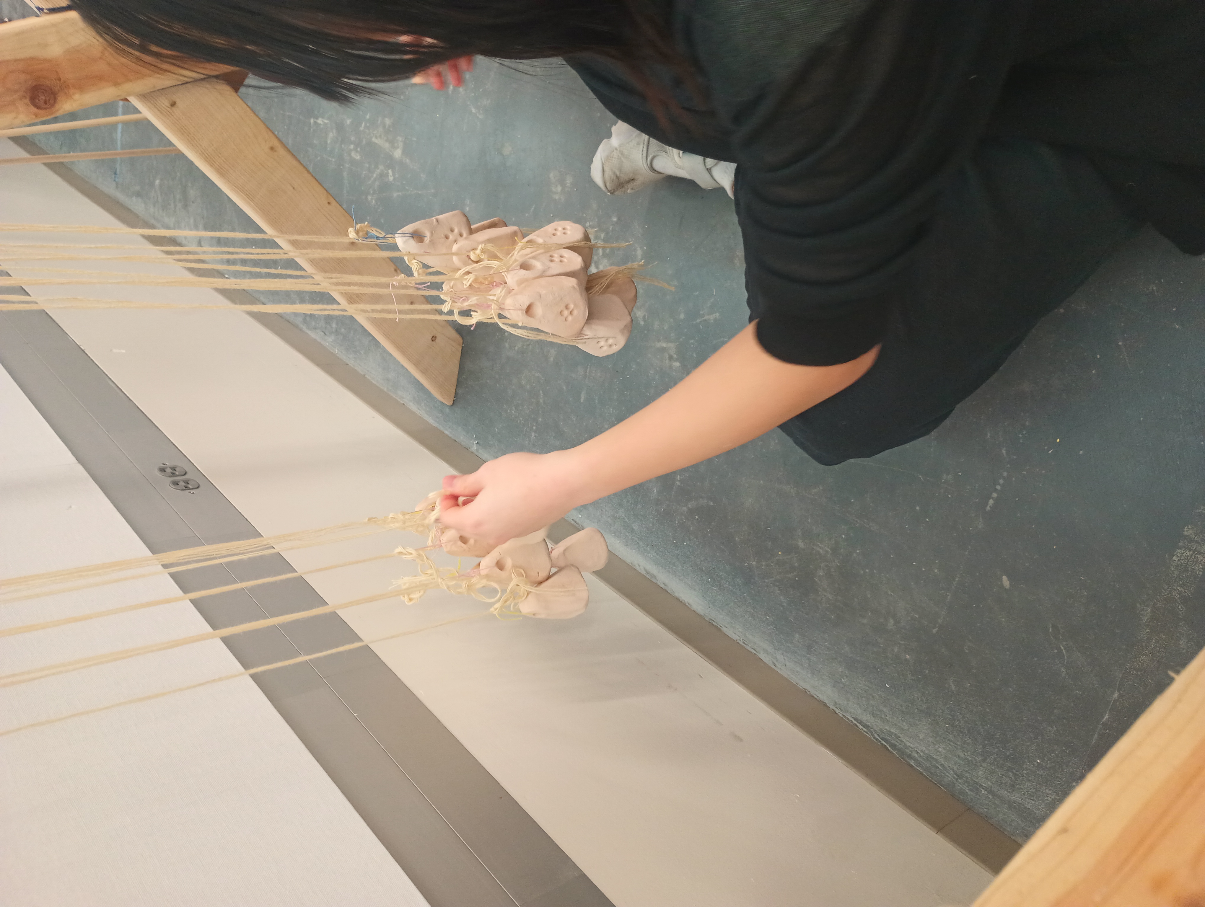 A student sits on the floor and ties a bundle of warp threads to a loom weight. Already there are two rows of loom weights tied to warp threads hanging. The loom weights are pyramidal and made of fired clay.