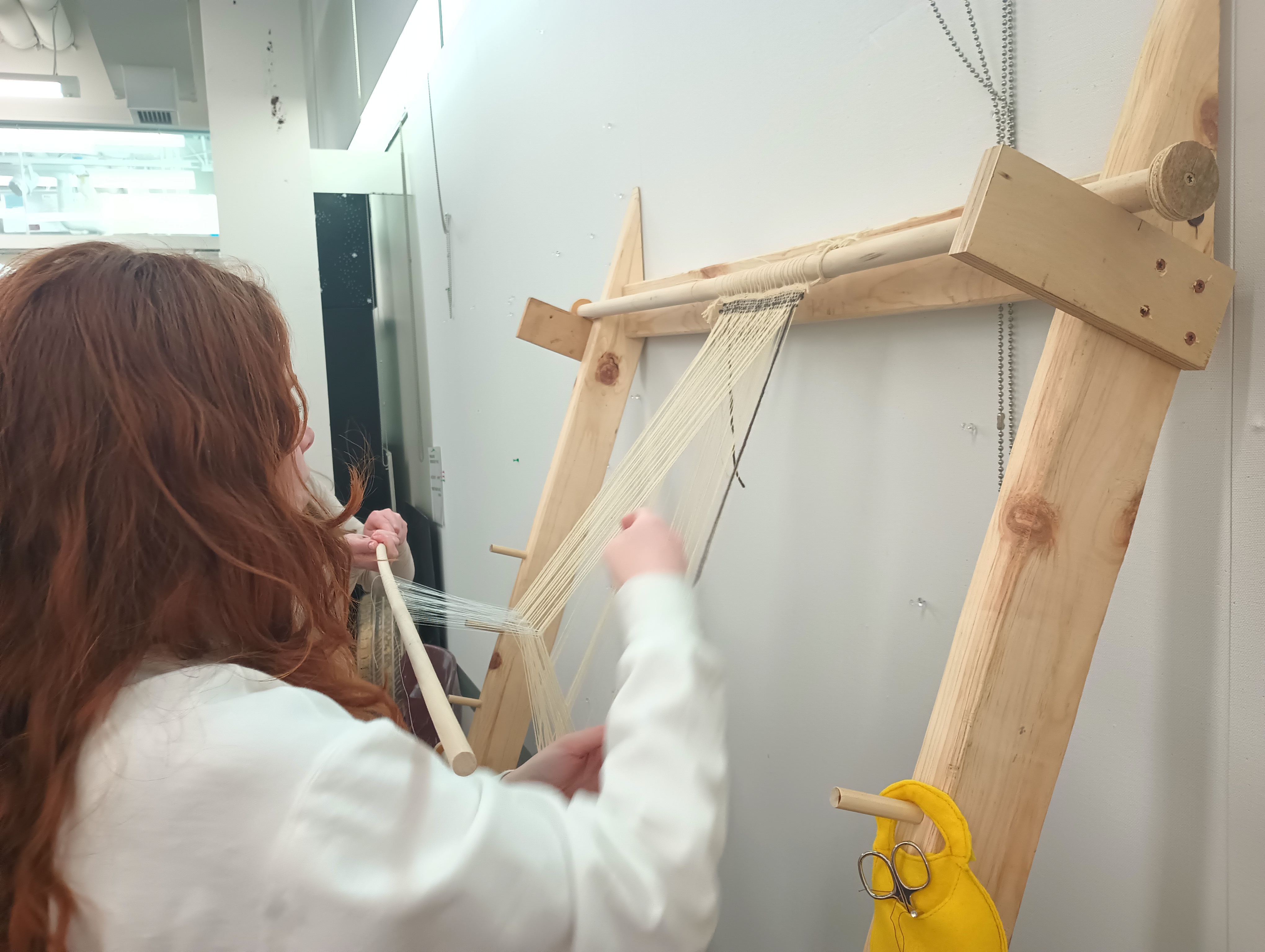 One student pulls the heddle bar forward. The string heddles pull every other warp thread forward, creating a shed through which a second student prepares to pass a bobbin of yarn.