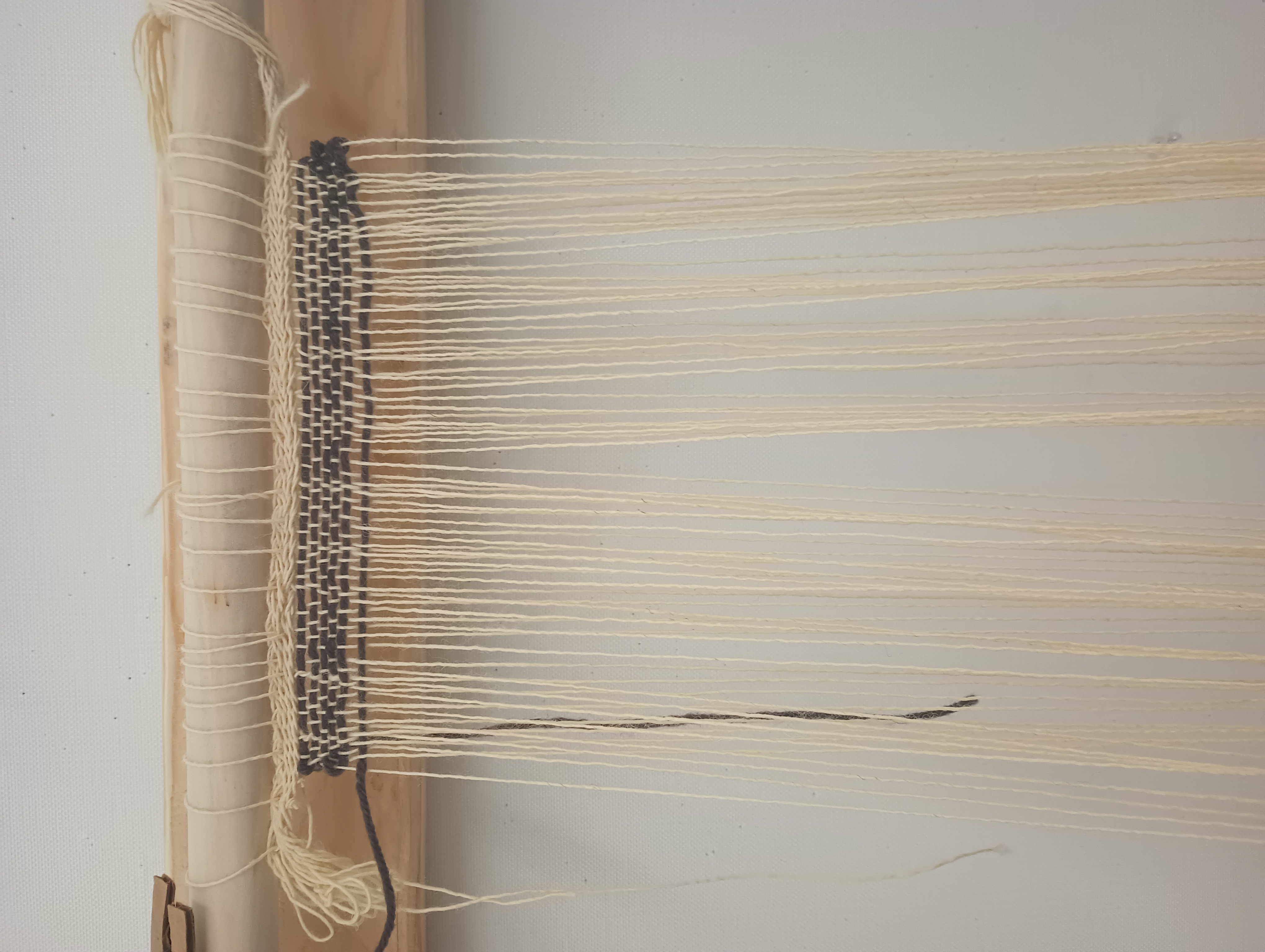 A close-up shot of the top of the warp-weighted loom showing the woven fabric. The warp threads are an undyed pale wool, and a thicker grey yarn has been woven in as weft.
