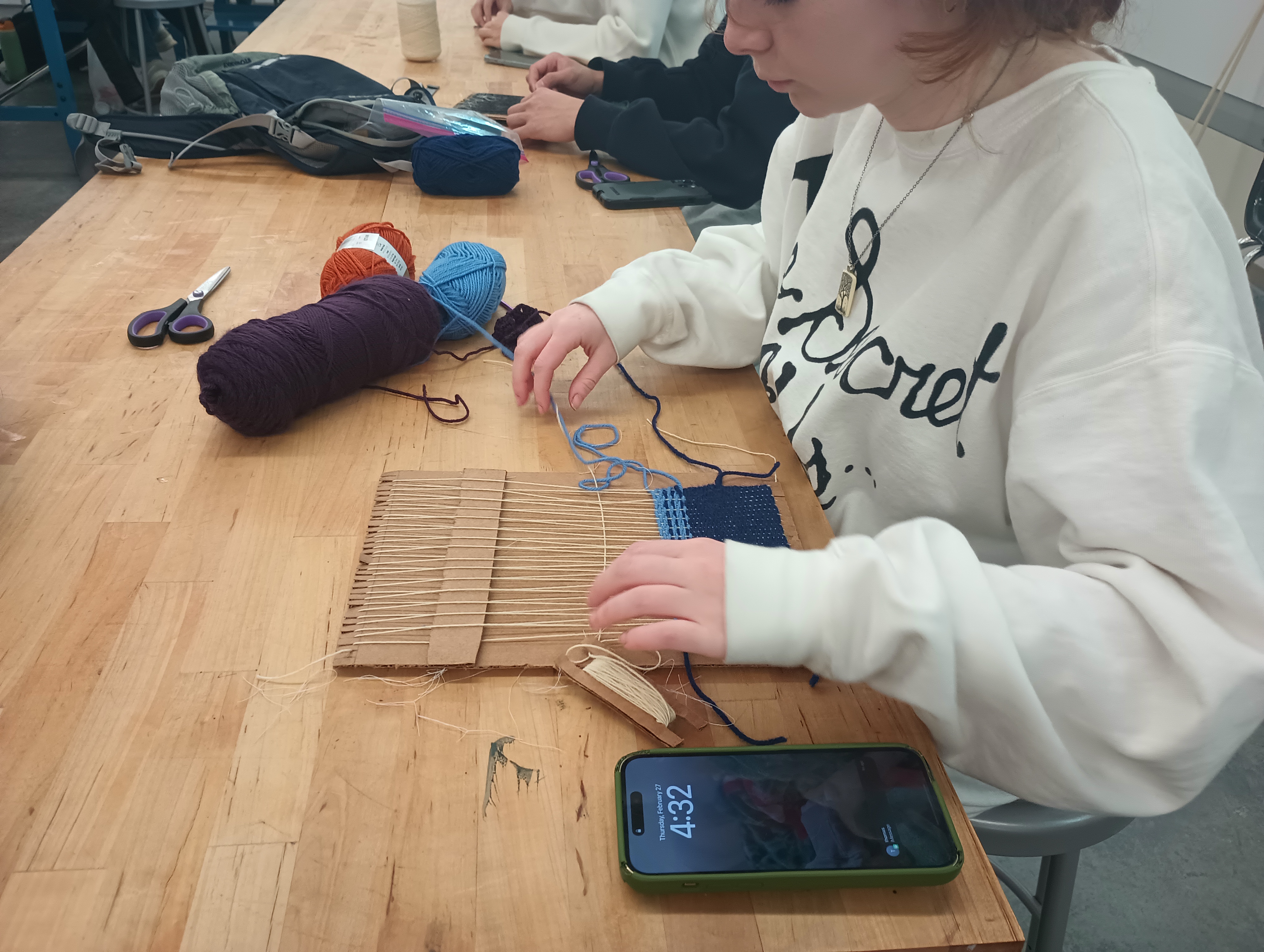 A student weaves a white weft on a small cardboard loom with a shed bar. The fabric she has already woven has stripes are different shades of blue.