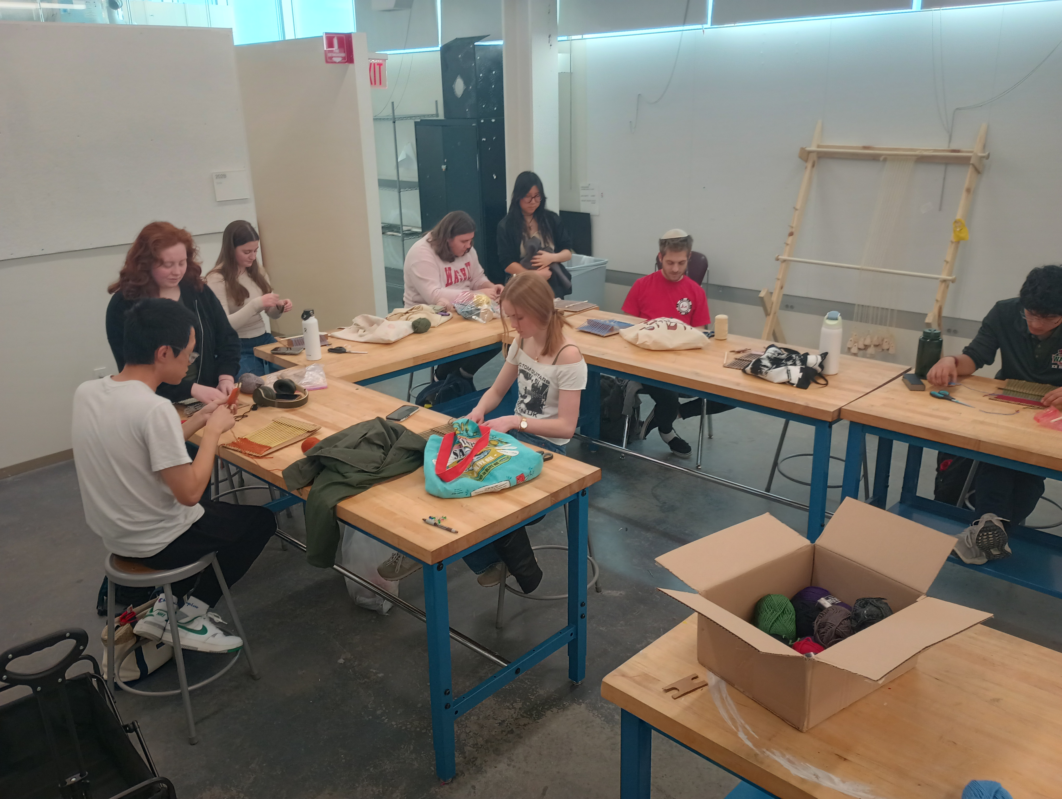 Students sit at tables in a classroom working on weaving rectangles on small cardboard looms. There is a warp-weighted loom leaning against the far wall of the classroom.